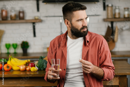 The guy with beard holds vitamins, pills and glass of water in his hand in the kitchen and glass of water. Medicine, health care, pharmacy and people concept.