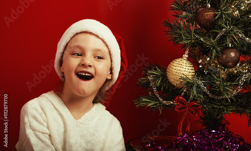 Fun emotional kid girl with laughing facial grimacing near the decor holiday fur green tree in red santa claus hat. Happy expression winter holadays with small natural emotion child closeup photo