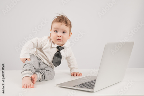 A cute child wearing a suit holds glasses and lies behind a laptop.