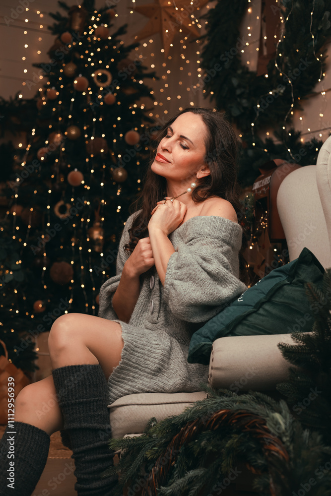 Woman sits by a beautifully decorated Christmas tree, wearing a cozy sweater. Warm lights and holiday ornaments, festive atmosphere