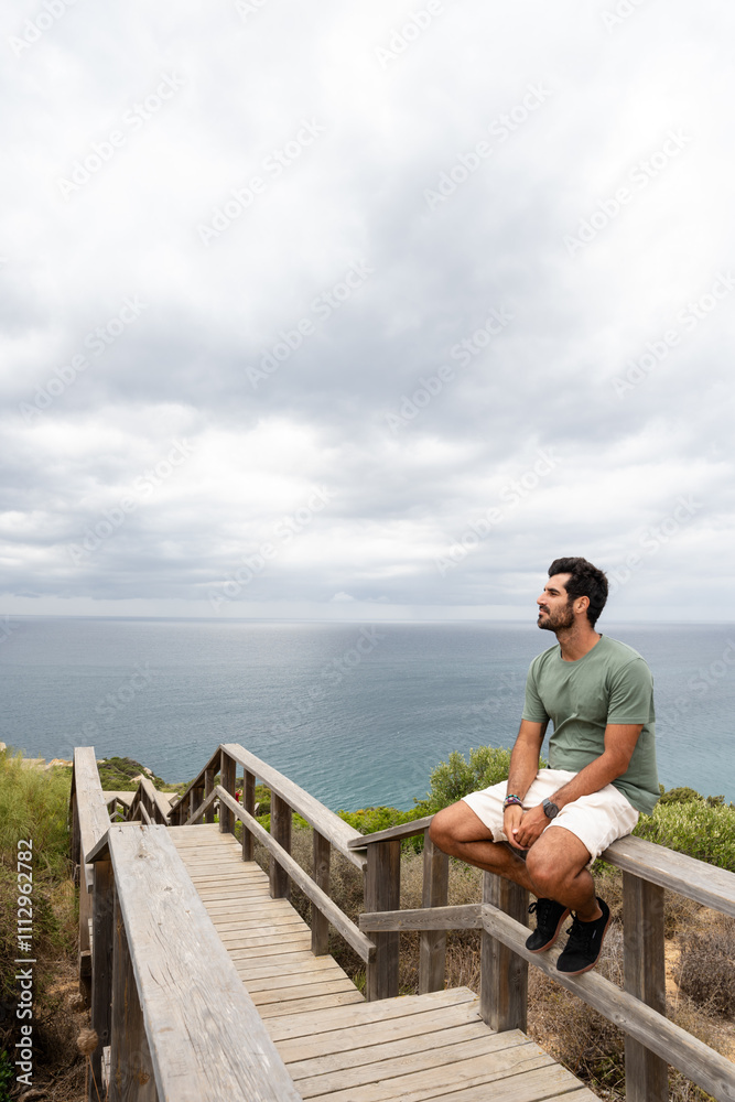 Young man sitting on wooden railing enjoying ocean view Young man sitting on wooden railing enjoying ocean view