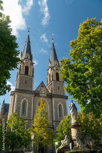 A majestic church with twin spires rises against a bright blue sky, surrounded by lush green trees and a decorative fountain featuring classical statues in the foreground. photo