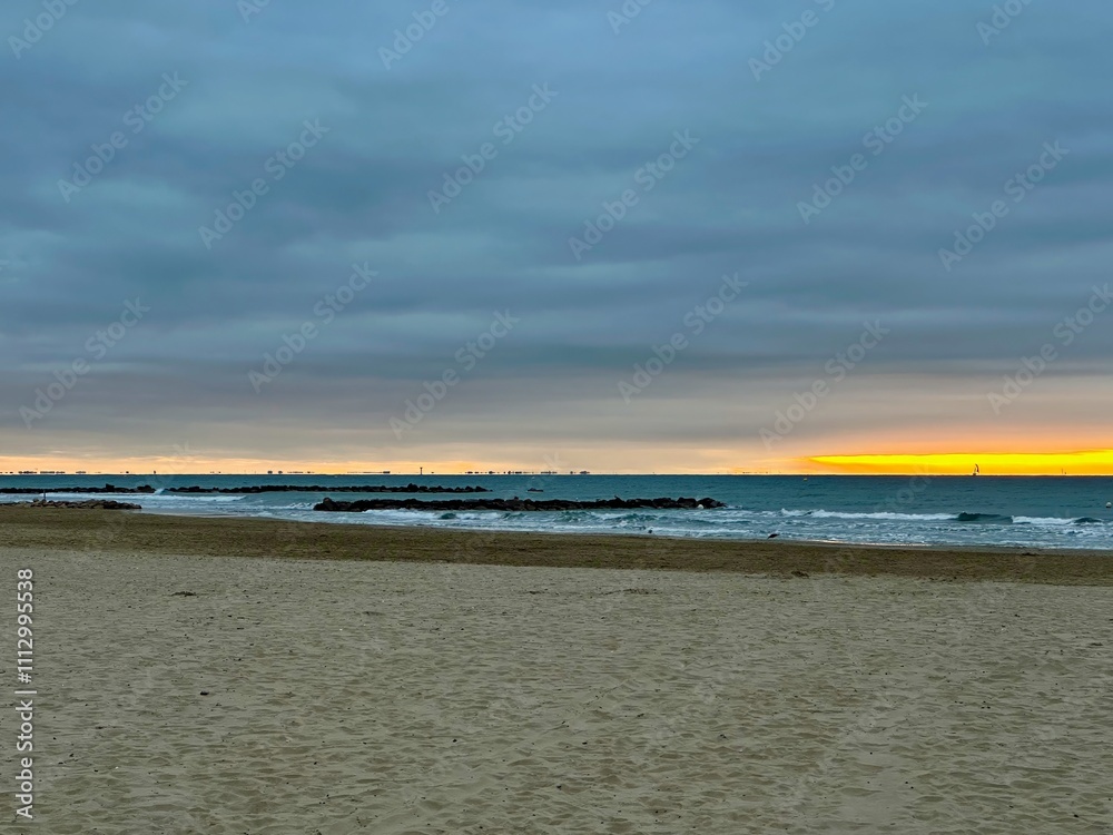 Obraz premium Golden Sunrise Over the Mediterranean Sea with Sailboat on the Horizon, Reflections of Grau-du-Roi and La Grande-Motte, Breakwaters, and Sandy Beach at Palavas-les-Flots