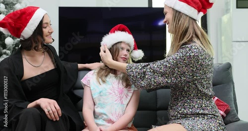 Mother puts Santa hats on daughter head sitting on sofa with aunt during Christmas Eve. Queer family with little girl celebrates New Year together