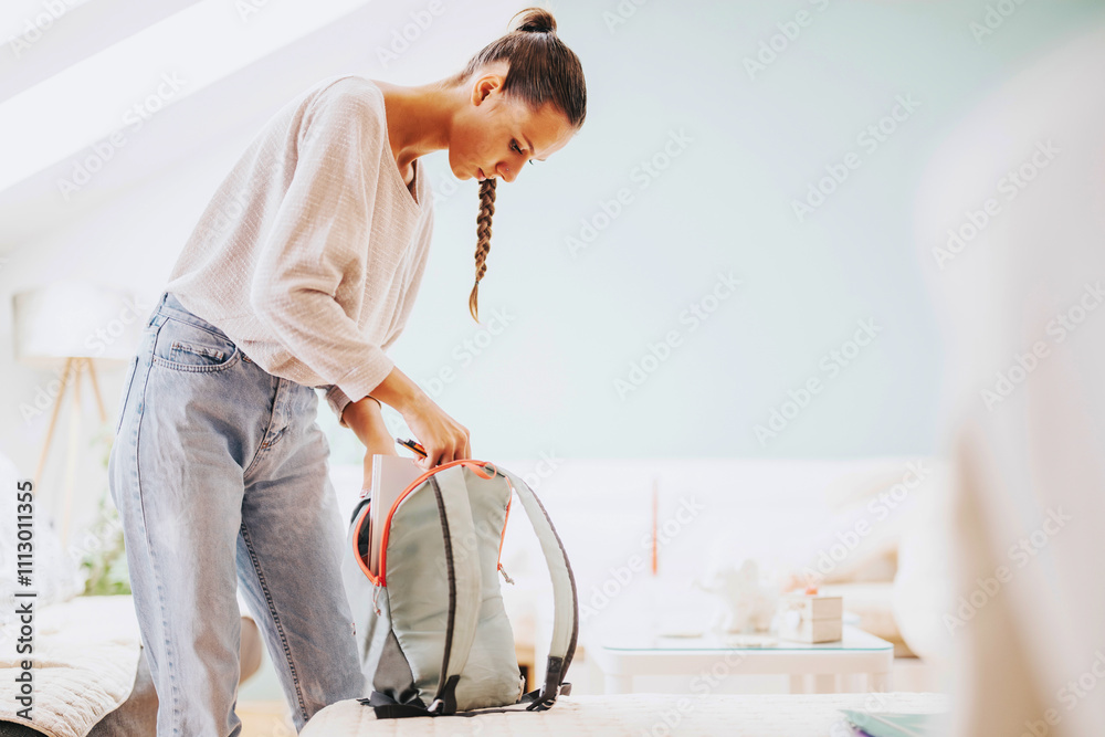 Fototapeta premium Young college student at home, preparing their backpack for a study session. The relaxed and tidy home environment enhances focus and comfort.
