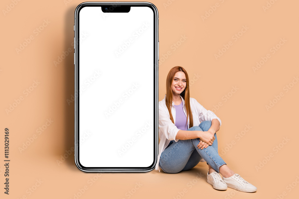 Full size photo of gorgeous woman wear white cardigan sitting near smartphone display empty space isolated on beige color background