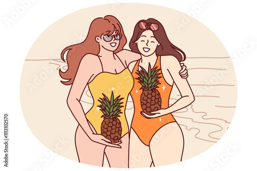 Two women in swimsuits stand on beach with pineapples in hands and smile inviting visit tropical island. Young happy girls in swimsuits spend holidays on sunny beach near sea or ocean