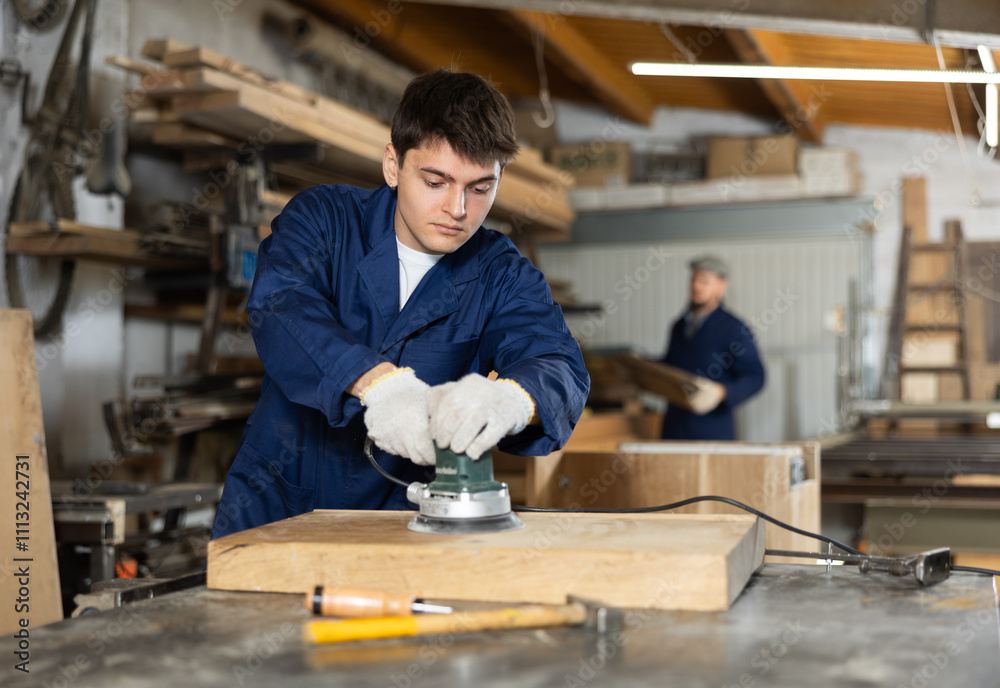Young guy worker in carpentry workshop polishes wooden board with machine Young guy worker in carpentry workshop polishes wooden board with machine