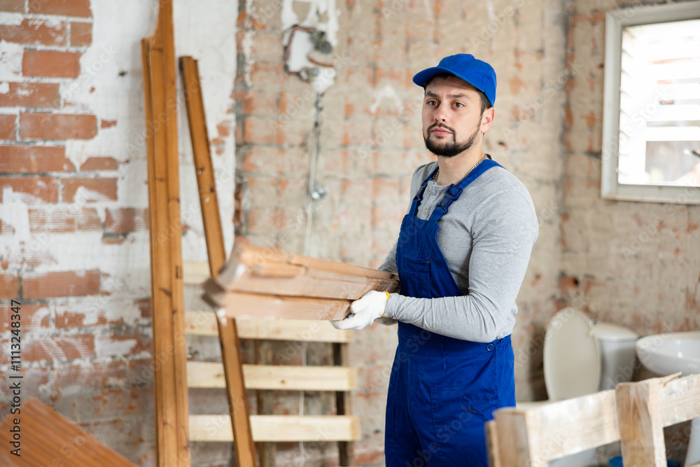 Young bearded builder in blue overalls preparing materials for work in building under renovation, arranging timber.. Young bearded builder in blue overalls preparing materials for work in building under renovation, arranging timber..