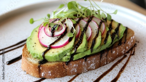 A close-up shot of a slice of avocado toast with sliced radish and balsamic glaze drizzled over the top. The toast is on a white plate with a speckled pattern. photo