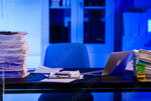 A quiet office desk at night, illuminated by a desk lamp. Documents, papers, and files are scattered across the desk, reflecting a late-night overtime work atmosphere.