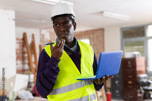 Portrait of african-american man builder in vest with laptop standing in constructin site and having telephone conversation. photo