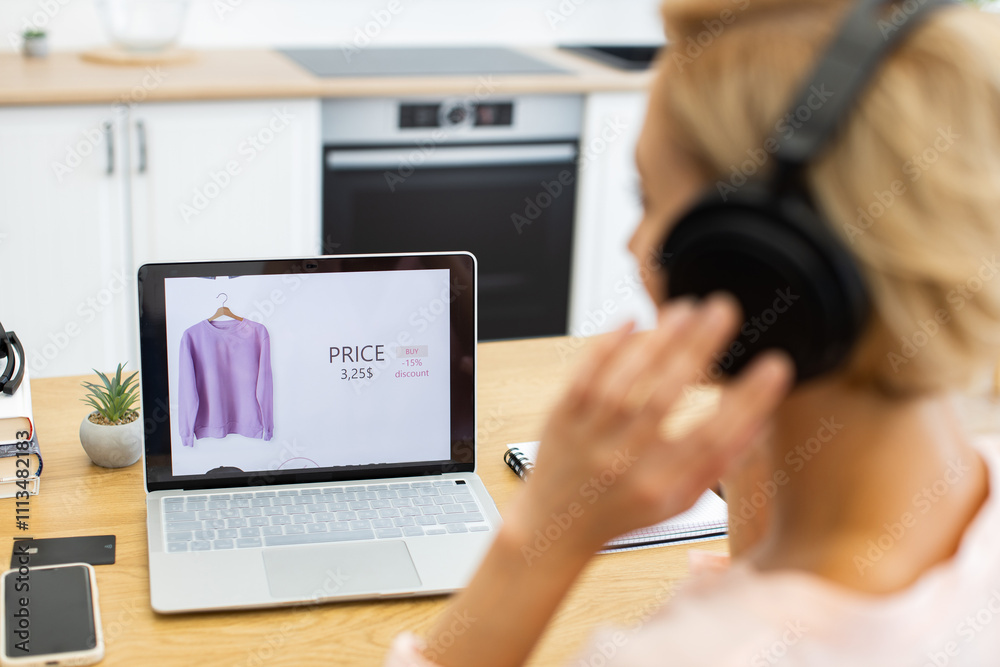 Happy Caucasian woman ordering clothes online using delivery website on laptop while listening to music with headphones. Image shows online shopping interface with discounted price. Happy Caucasian woman ordering clothes online using delivery website on laptop while listening to music with headphones. Image shows online shopping interface with discounted price.