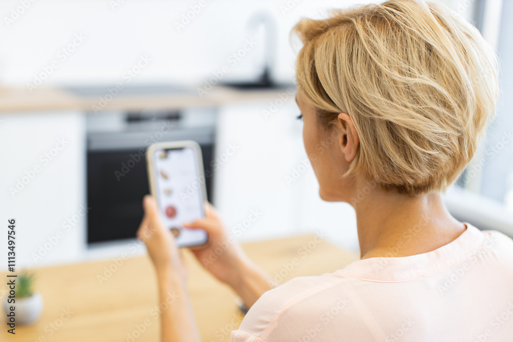 Caucasian woman using smartphone to order food online in modern kitchen setting. Blonde woman with short hair browsing delivery website. Image reflects convenience, technology, and digital lifestyle. Caucasian woman using smartphone to order food online in modern kitchen setting. Blonde woman with short hair browsing delivery website. Image reflects convenience, technology, and digital lifestyle.