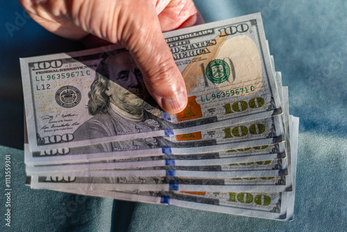 Close-up of a person's hand holding a fan of 100 American dollar banknotes against green fabric background. Photo taken outdoors with sun light and shadows.