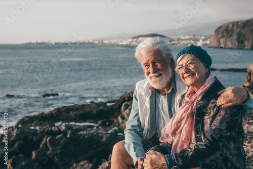 Cheerful lovely senior couple sitting embracing on the rocks at sea enjoying sunset light. Relaxed lifestyle for a caucasian couple in vacation or retirement photo