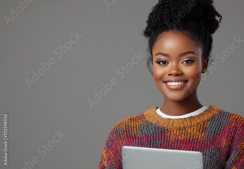 Young woman smiling while holding a laptop in a cozy, stylish sweater against...