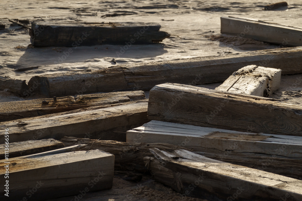 Weathered wooden beams lie scattered on sandy shore as sun casts warm golden light. Texture of aged wood contrasts with smooth sand, creating a captivating natural composition of textures and colors Weathered wooden beams lie scattered on sandy shore as sun casts warm golden light. Texture of aged wood contrasts with smooth sand, creating a captivating natural composition of textures and colors