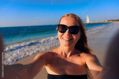 Beautiful woman takes selfies during a summer holiday at the sea. Attractive caucasian woman in a swimsuit makes a selfie on a smartphone while walking at the sunrise by the sea.