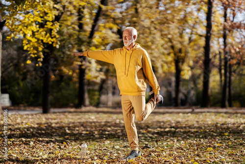 An old fit athlete in tracksuit balancing on one foot and stretching it while preparing for fitness in nature.