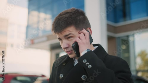 Close up of man in black coat talking on two phones interchangeably with focused expression in vibrant urban area featuring modern glass buildings photo