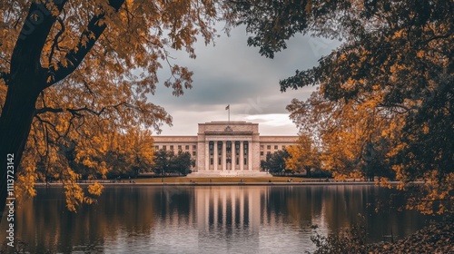 The Federal Reserve building stands prominently near a tranquil pond, framed by vibrant autumn foliage, reflecting a calm, cloudy sky overhead.
