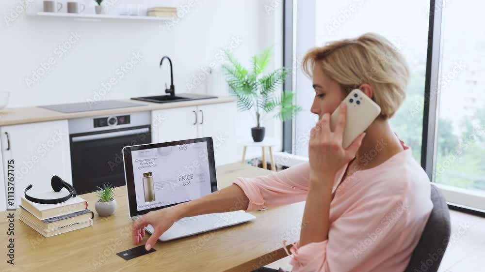 Caucasian woman ordering cosmetics online using laptop while talking on phone and holding credit card. Blonde hair, adult female engaged in online shopping and payment in bright home environment.