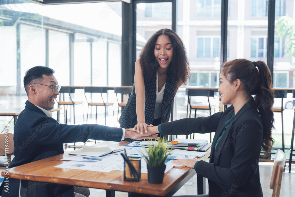 © Kritdanai - United in Success: A diverse team of business professionals in a modern office setting, their hands stacked together in a gesture of unity and collaboration. They are excited and happy.
