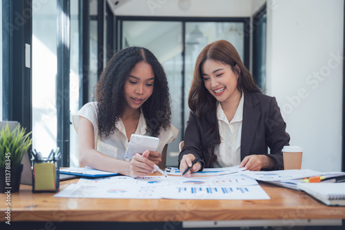 Collaborative Businesswomen Analyzing Data: Two smiling businesswomen, one African American and one Asian, collaborate closely, reviewing financial charts and graphs.