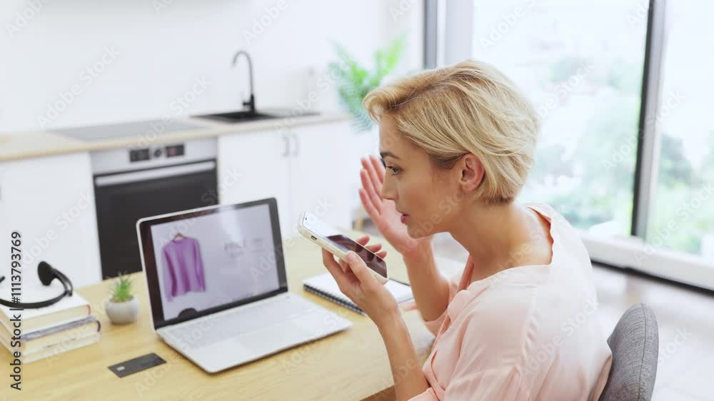 Caucasian woman talking on phone expressing concern about clothing purchase from online store. Middle-aged female sitting in home office with laptop displaying website. Caucasian woman talking on phone expressing concern about clothing purchase from online store. Middle-aged female sitting in home office with laptop displaying website.