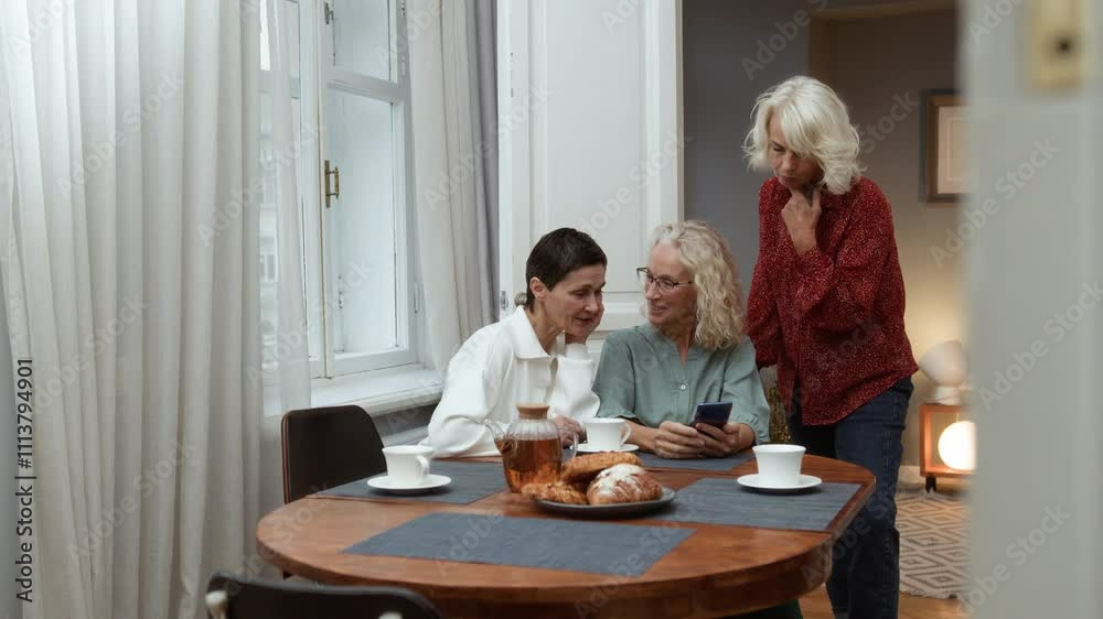 Three elderly women sit at table chatting joyfully, looking in mobile telephone. Elegance blonde senior women chill together talking. Female aged friends have tea time at home and communicate smiling Three elderly women sit at table chatting joyfully, looking in mobile telephone. Elegance blonde senior women chill together talking. Female aged friends have tea time at home and communicate smiling