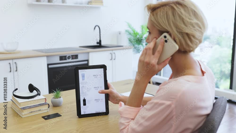 Caucasian woman using smartphone to order cosmetics online while browsing website on tablet. Seated at modern office desk, she multitasks efficiently in bright, stylish interior.