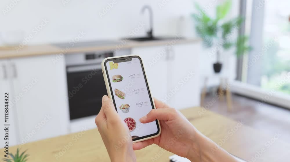 Caucasian woman using smartphone for online food order in bright modern kitchen. Person browsing delivery app menu with various meal options like sandwiches and juices.