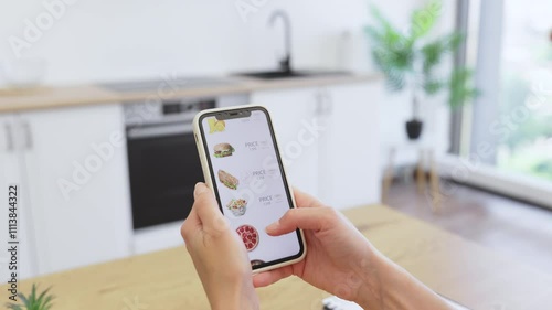 Caucasian woman using smartphone for online food order in bright modern kitchen. Person browsing delivery app menu with various meal options like sandwiches and juices. photo