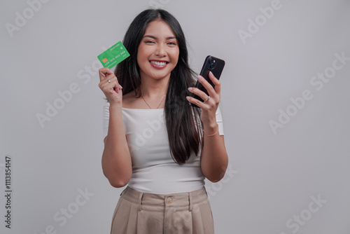 Excited young Asian woman showing a credit card and smartphone she holds.