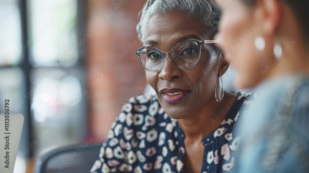 Woman with glasses is talking to another woman. The woman with glasses is wearing a blue floral dress Woman with glasses is talking to another woman. The woman with glasses is wearing a blue floral dress