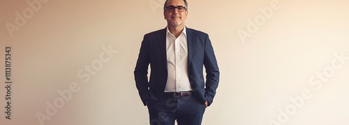 A man in his late 40s wearing a dark blue suit and glasses, standing with his hands in his pockets against a soft beige background with lots of empty space.
