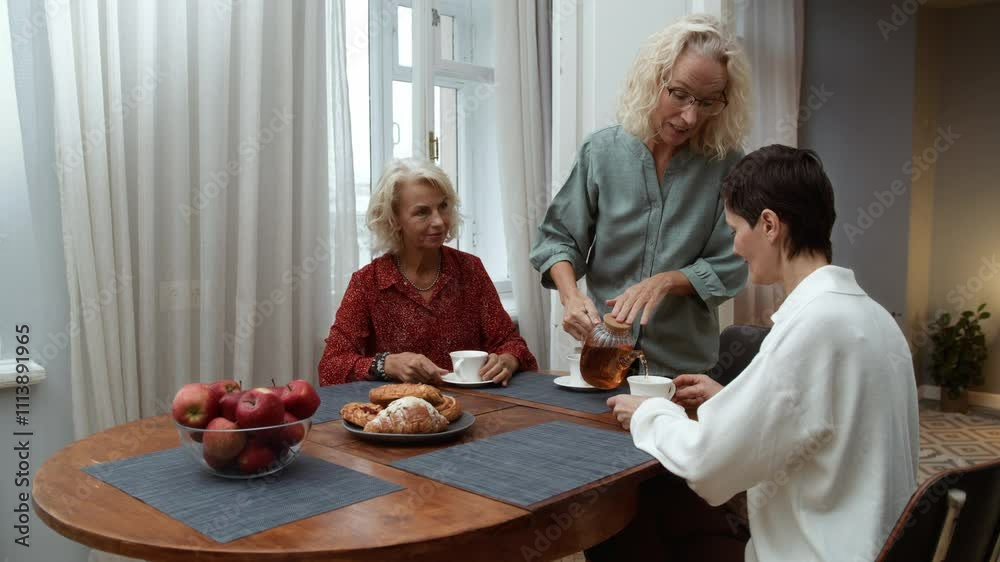 Three elderly women sitting at table chatting joyfully. Elegance blonde senior women chill together talking. Female aged friends having tea time at home and communicating smiling Three elderly women sitting at table chatting joyfully. Elegance blonde senior women chill together talking. Female aged friends having tea time at home and communicating smiling