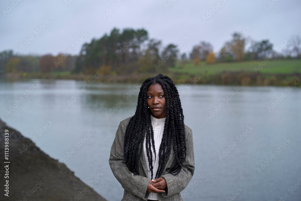 Woman with long braided hair wearing a gray coat stands near a tran0uil lake on an overcast day, Brandenburg, Germany Woman with long braided hair wearing a gray coat stands near a tran0uil lake on an overcast day, Brandenburg, Germany
