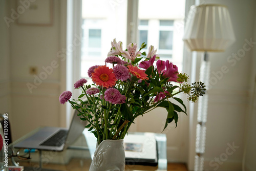 Fresh colorful flowers in a white vase on a table with a laptop and lamp in a bright room. photo