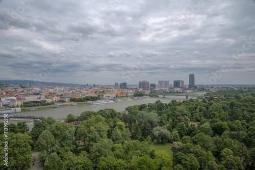 Elevated view of Bratislava’s urban skyline, the Danube River, and surrounding greenery under a cloudy sky.