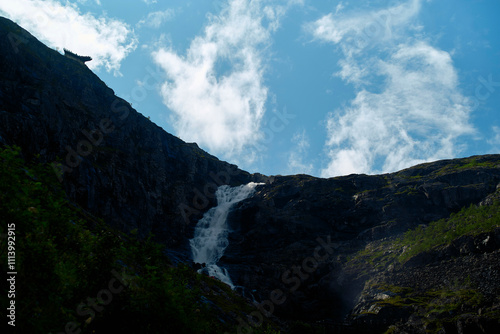 A cascading waterfall flows down a rugged cliffside under a blue sky speckled with white clouds, Trollstiege, Trollvegen, Norway photo