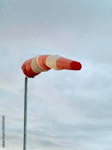 A red and white windsock fully extended against a cloudy sky, indicating strong wind conditions, Norway