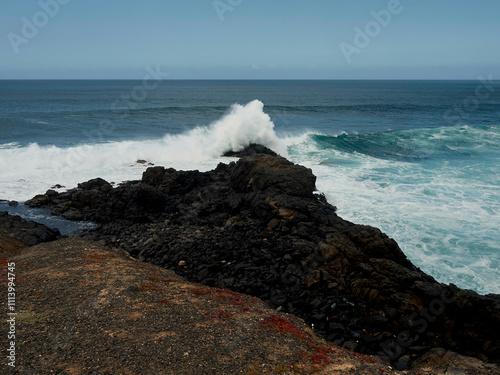 Waves crash against the rugged black rocks on a coastline under a cloudy sky, Fuerteventura, Spain photo