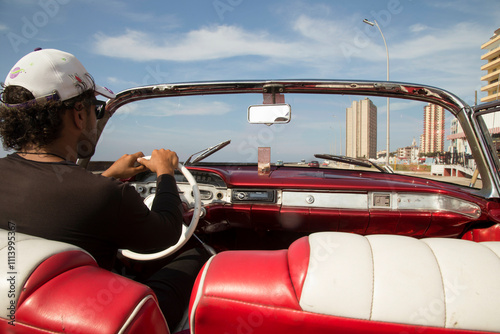 Man driving a vintage convertible car with a view of a cityscape in the background, Havana, Cuba photo