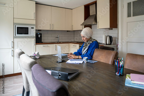 Woman in a hijab concentrating on a laptop at a kitchen table with papers and stationery around her. photo