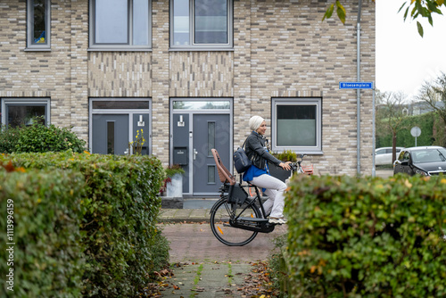 Woman riding a bicycle on a residential street next to a brick house and parked cars under an overcast sky. photo