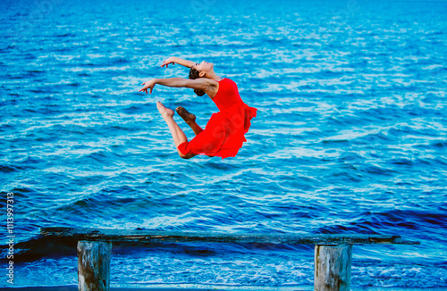 Graceful ballet dancer in a red dress performing a jump over blue water with a wooden structure in the foreground, USA photo