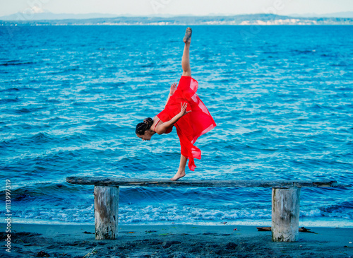 Young gymnast performing a handstand on a wooden beam by the sea in a red dress, USA photo