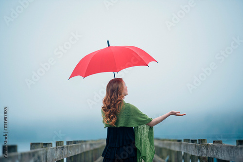 Woman with red umbrella standing on a wooden bridge extending her hand to feel the weather. photo